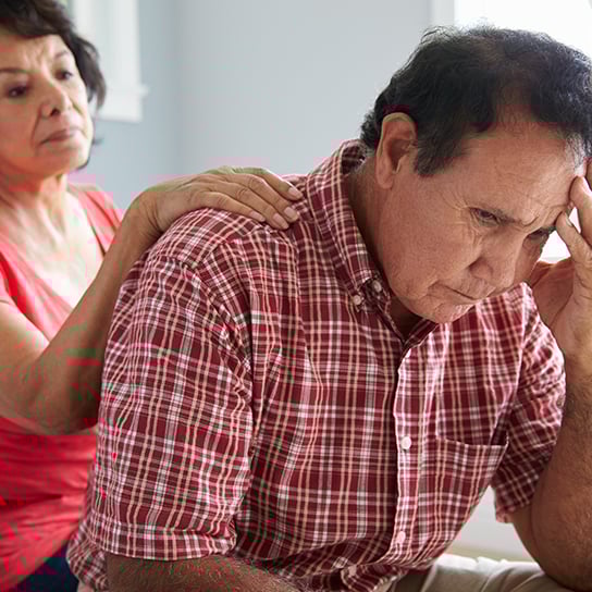Older man with head in his hand, with a woman sitting behind him with her hand on his shoulder