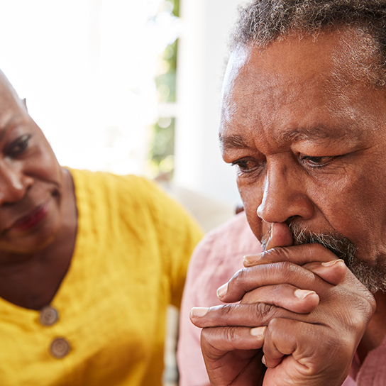 An older man with his hands folded in front of his mouth, with an older woman in the background, seemingly comforting him